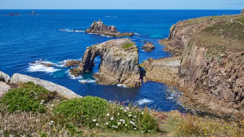 This landscape photograph shows the Enys Dodnan Arch and the surrounding cliffs on the coast of Cornwall, England, in the United Kingdom. The image was captured close to midday in summer, with the sunlight illuminating the blue sea and highlighting the rocky formations. The Enys Dodnan Arch, a natural rock structure, is prominently situated near the cliffs, and beyond it, the Longships Lighthouse can be seen on the horizon. Coastal vegetation and wildflowers are visible in the foreground, enhancing the view of the rugged shoreline in this region of Cornwall.